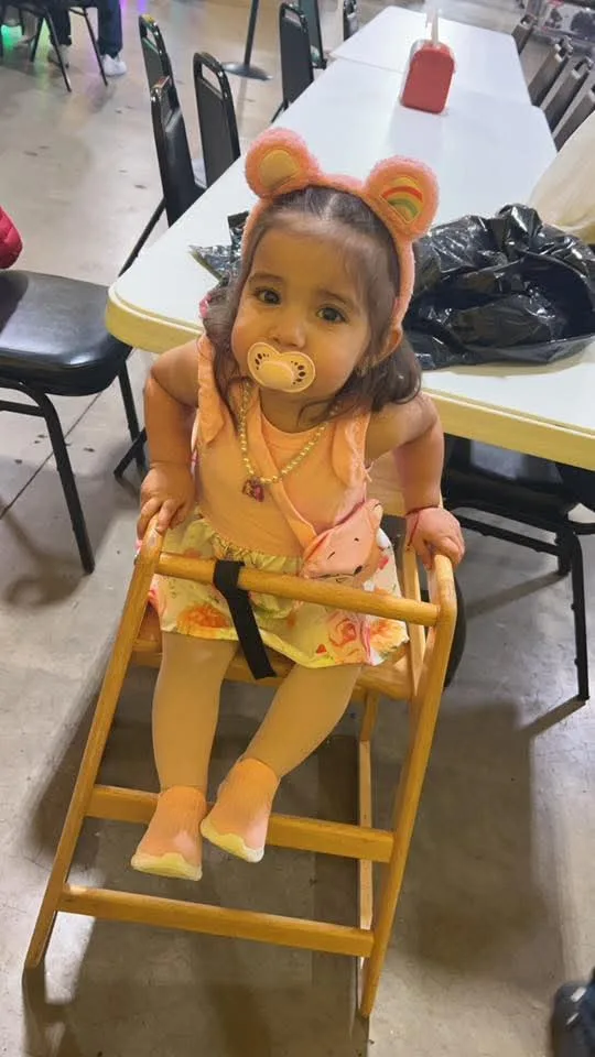 A toddler in a peach bear-ear headband sits in a wooden high chair at her birthday party at People’s Choice Family Fun Center, 2023.